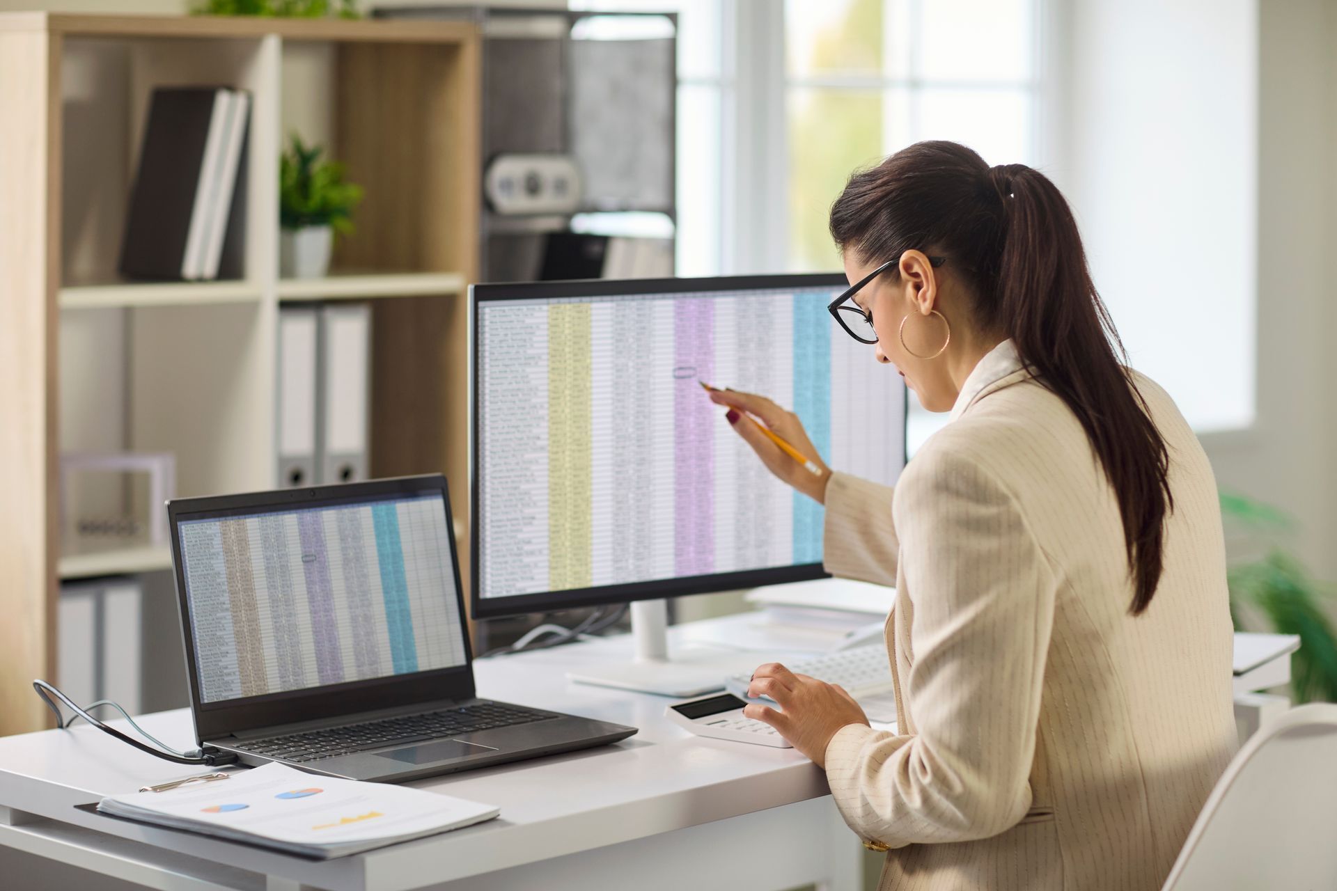 Woman working at a desk with a laptop and monitor, pointing at a spreadsheet in a bright office