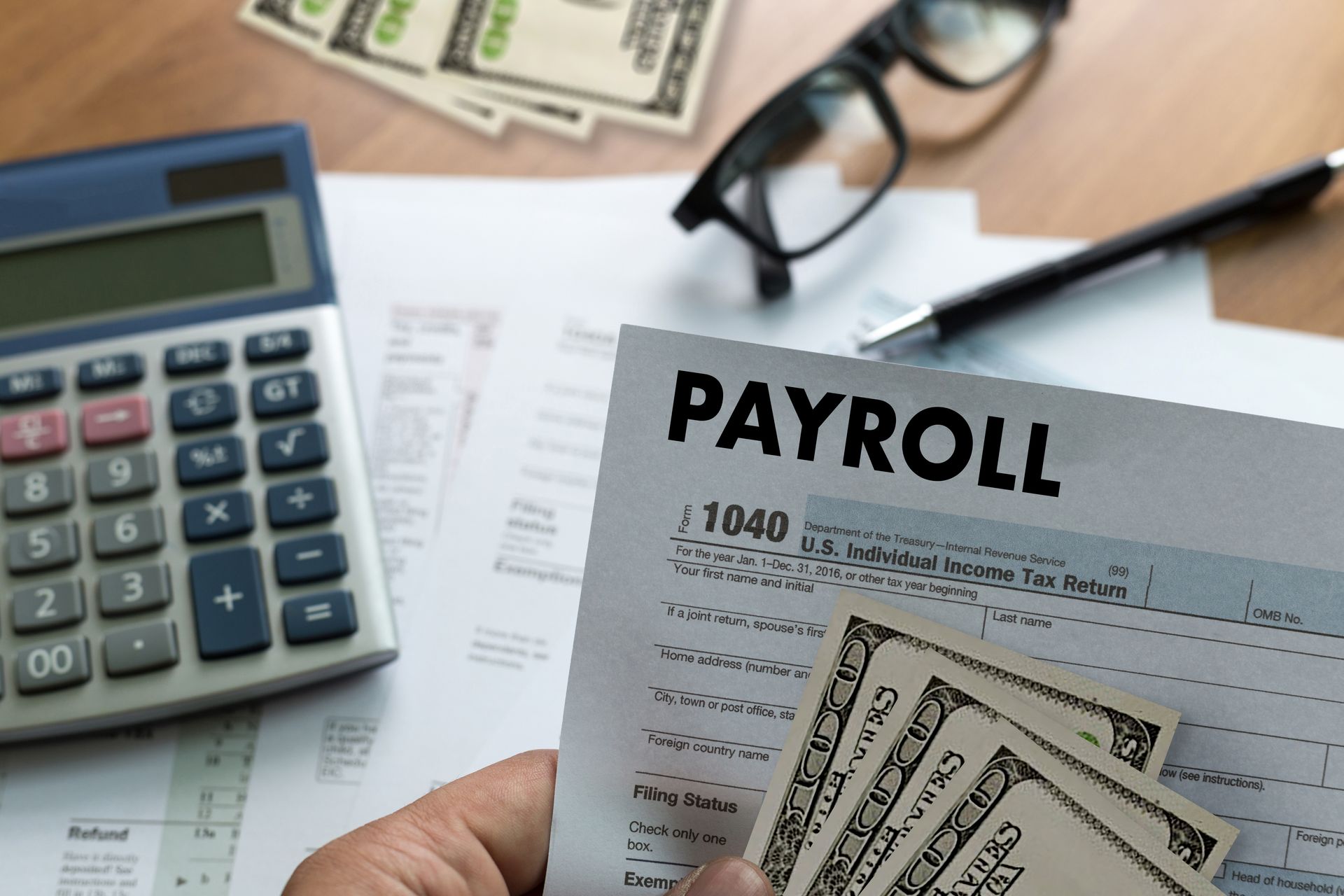 Payroll documents, calculator, glasses, and pen on a desk with cash and a 1040 tax form.