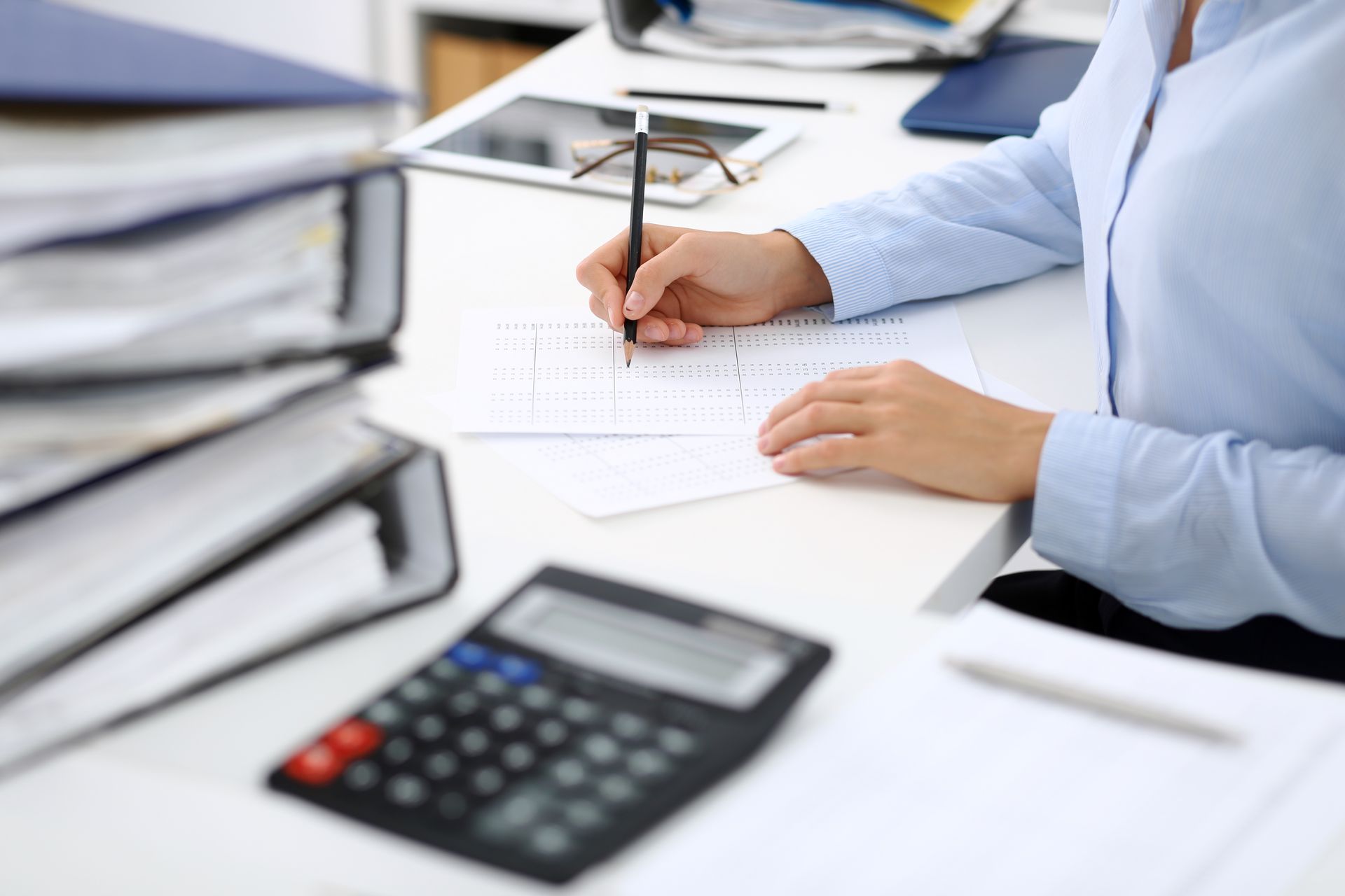 Person writing on a document at a desk with a calculator and stacked papers nearby