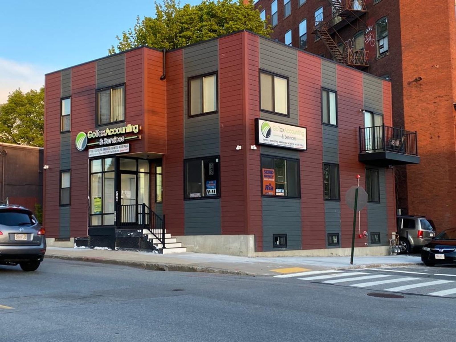 Brick storefront on a sunny street corner beside a crosswalk and parked cars