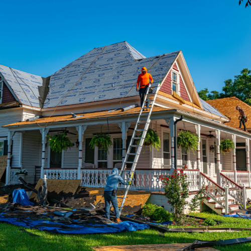 Two roofers on a ladder, working on a house roof. Blue tarp on the lawn. Sunny day.