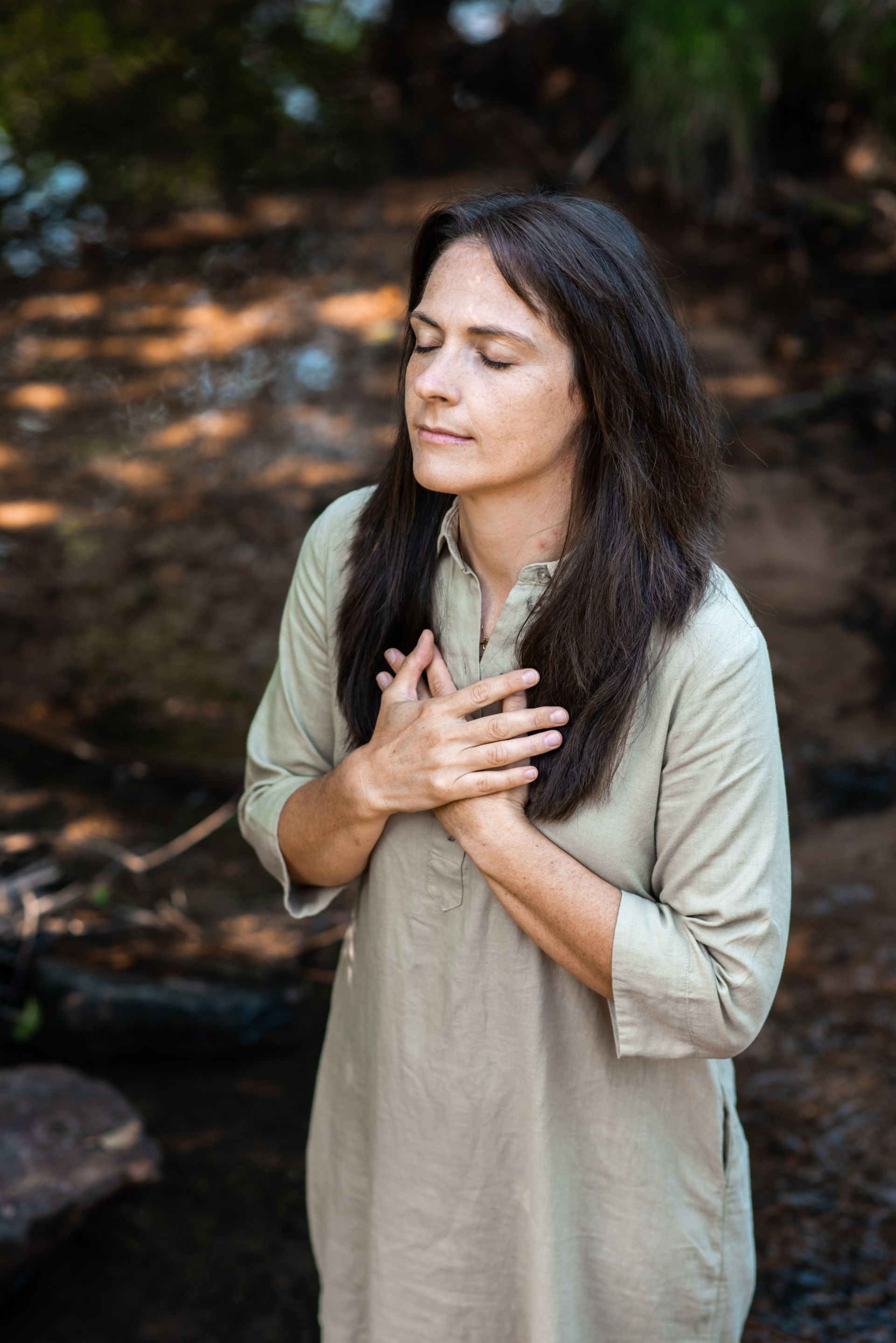Frau steht barfuß in einem Bach, Augen geschlossen, Hände auf der Brust. Trägt ein grünes Kleid, umgeben von Natur.