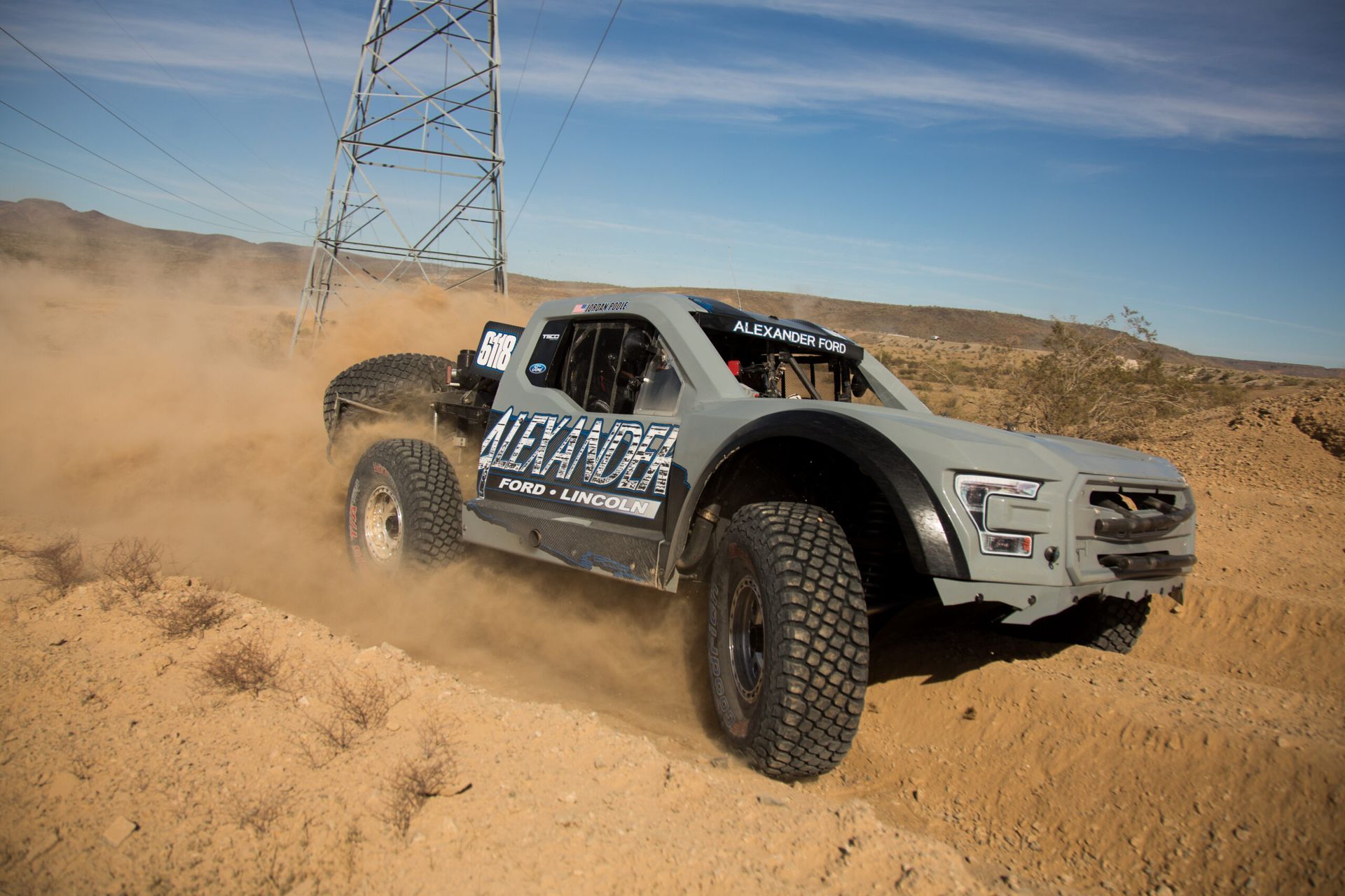 A gray truck is driving down a dirt road in the desert.