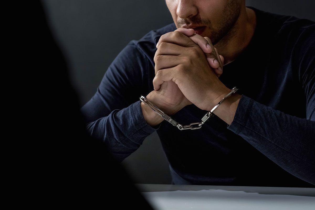 Man in handcuffs sits at a table, hands clasped, facing a person out of frame.