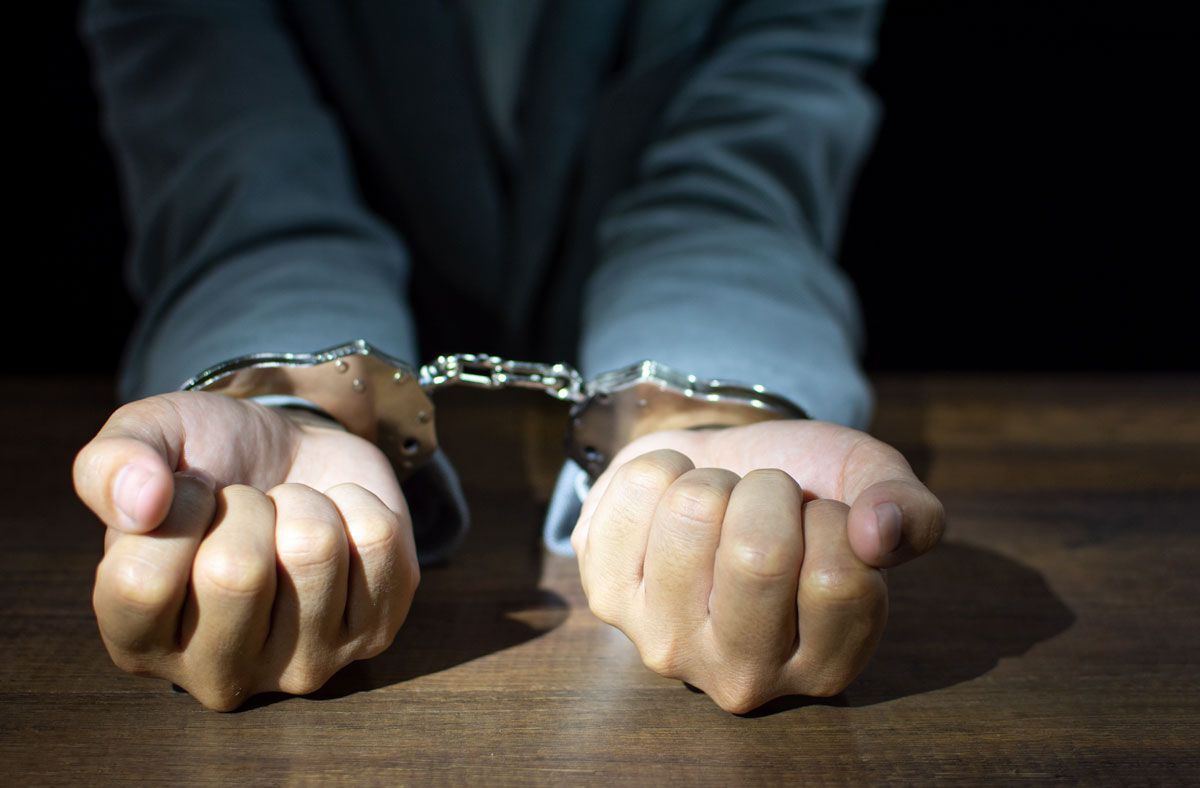 Hands in handcuffs on a wooden surface. Person in a suit, dark background.
