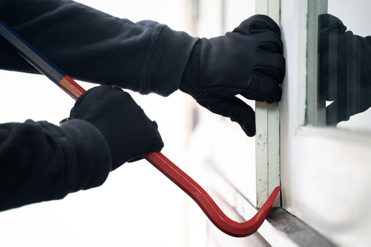 Person wearing black gloves using a red crowbar to pry open a window frame.