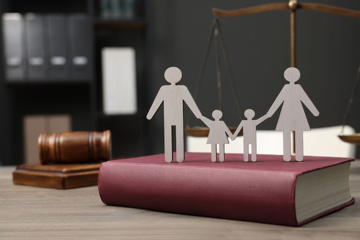 Paper family holding hands atop a law book near a scale and gavel, symbolizing family law.