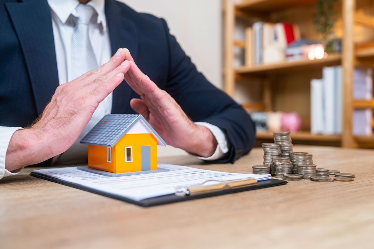 Person in suit protects model house with hands, next to money and documents.