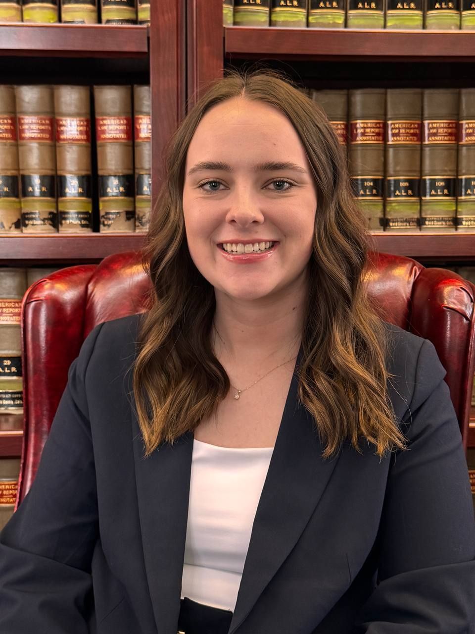 A person smiling, wearing a navy blazer and white top, seated in front of a bookshelf filled with law books.