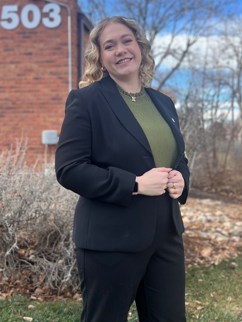 Young woman with blonde hair, smiling, wearing a black blazer and blouse, standing near a window.