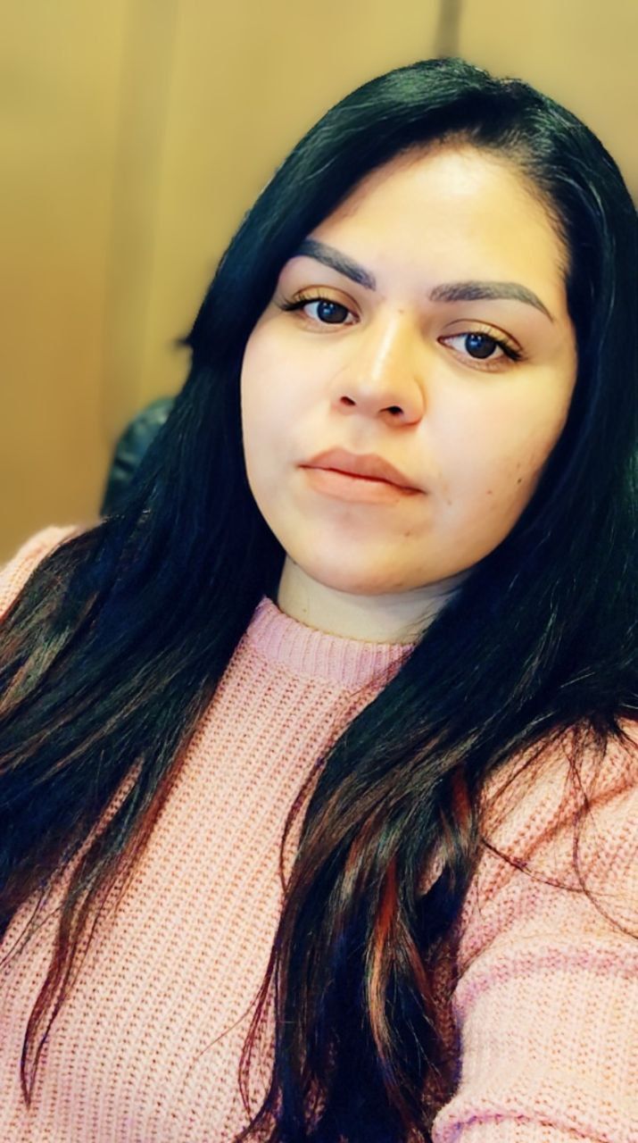 A person with long, dark hair wearing a light pink ribbed sweater, looking directly at the camera with a neutral expression.