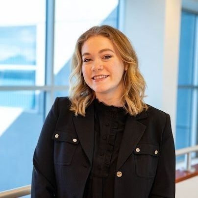 Young woman with blonde hair, smiling, wearing a black blazer and blouse, standing near a window.