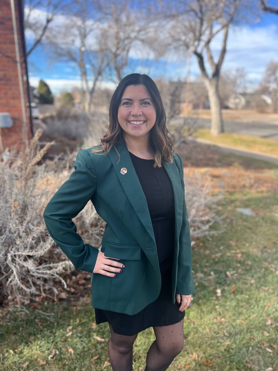 A smiling person wearing a dark green blazer over a black outfit, standing outdoors near bushes and trees.