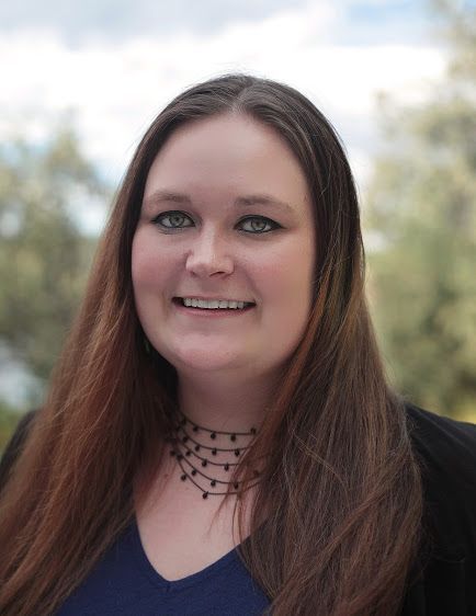 A person with long brown hair smiling at the camera, wearing a dark blue top and a multi-strand beaded necklace.