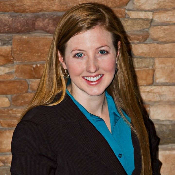 Woman with long auburn hair, smiling, wearing a blue shirt and black blazer, in front of a brick wall.