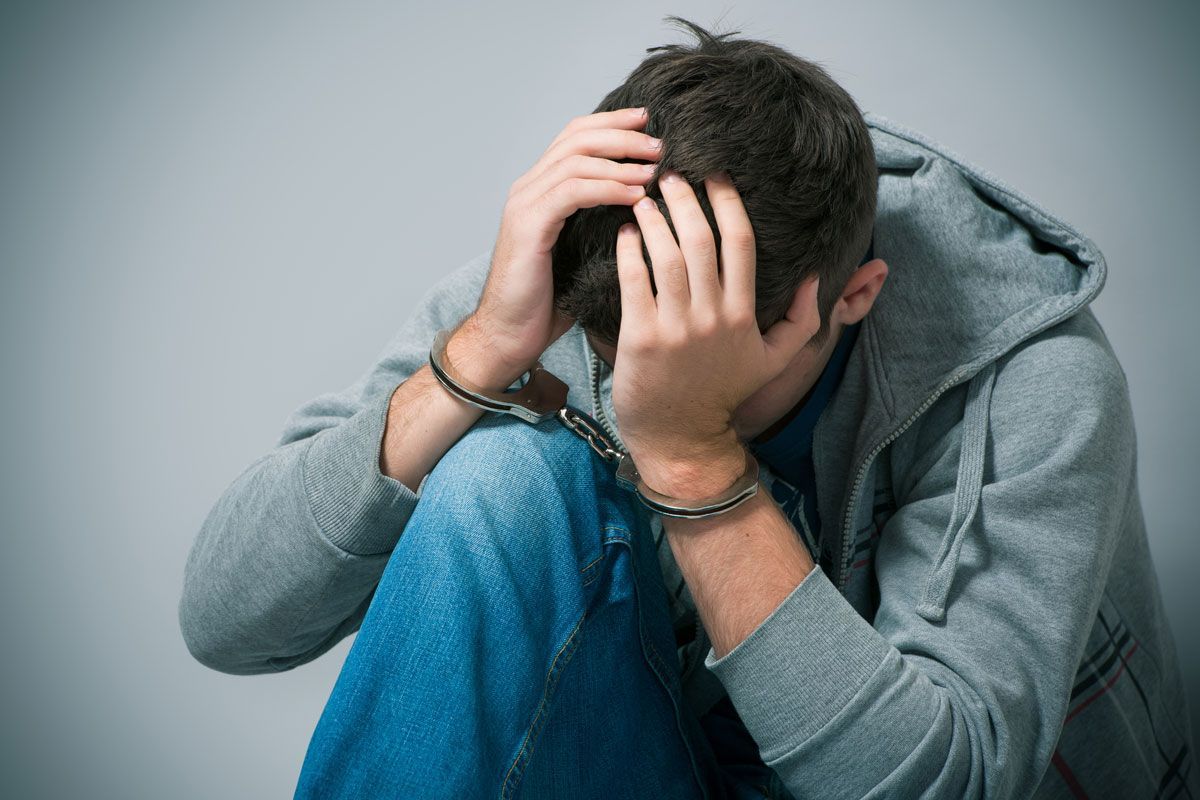 Man in handcuffs, head in hands, wearing hoodie and jeans, looking distressed.