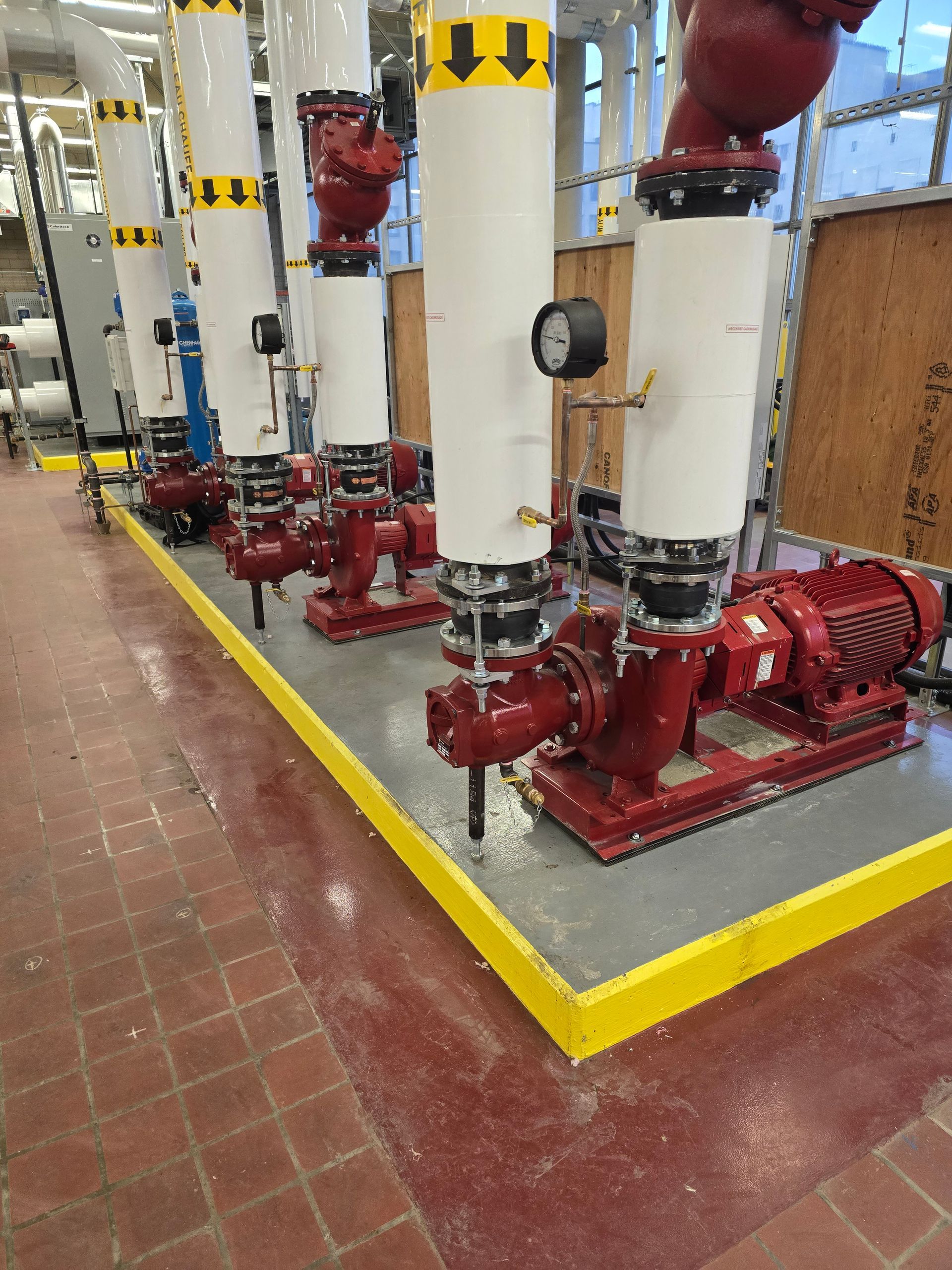Pompes rouges à tuyaux blancs et marquages ​​jaunes dans un local technique au sol carrelé de rouge.