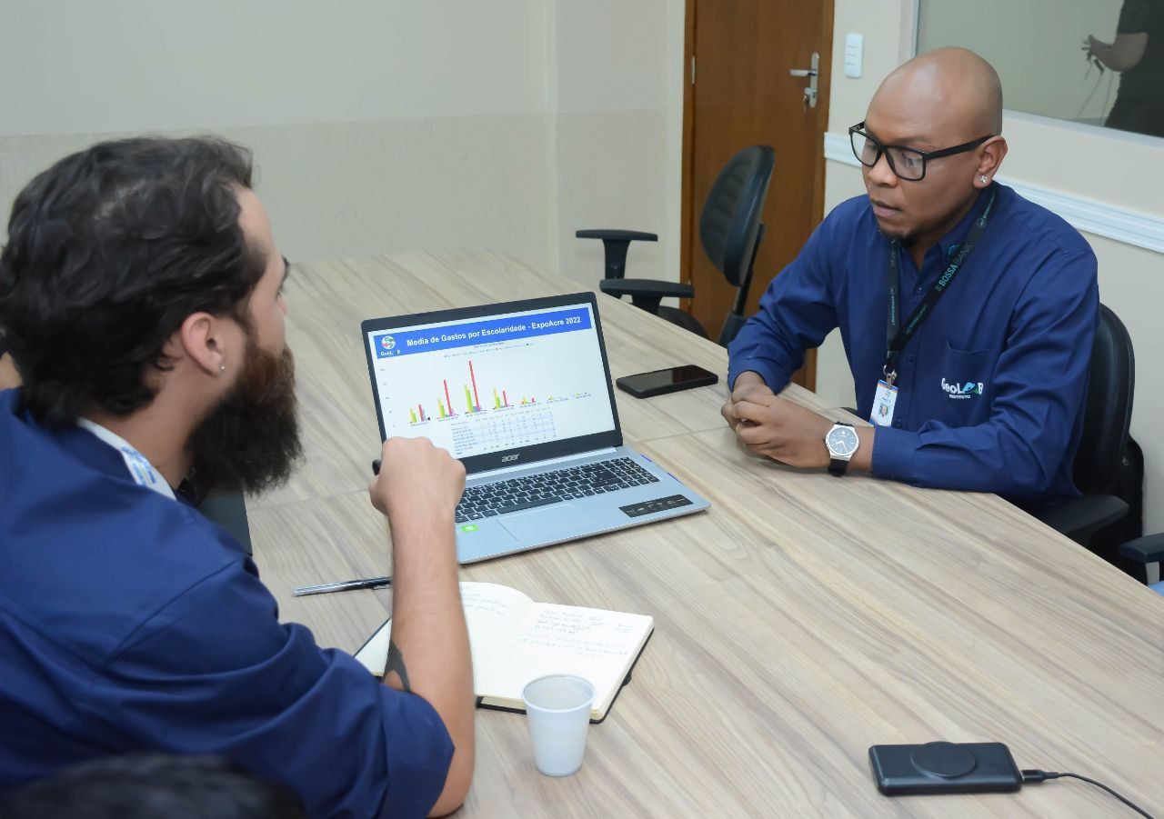 Dois homens estão sentados em uma mesa com um laptop sobre ela.
