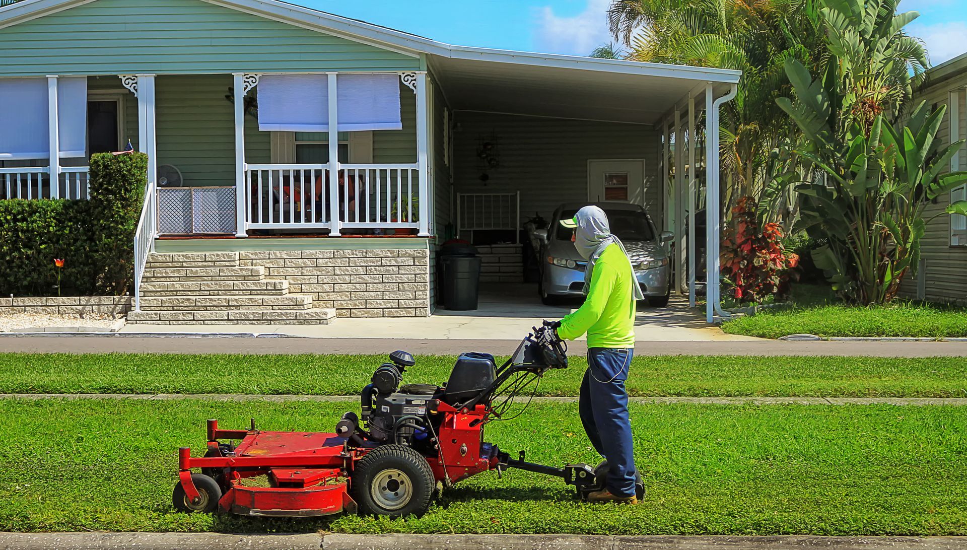 A worker in a bright green shirt mows a lawn in front of a house with a white porch and carport.