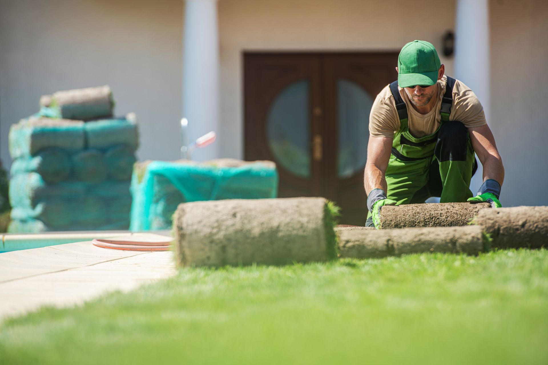 A worker in green workwear and a cap lays down a roll of sod on a residential lawn.