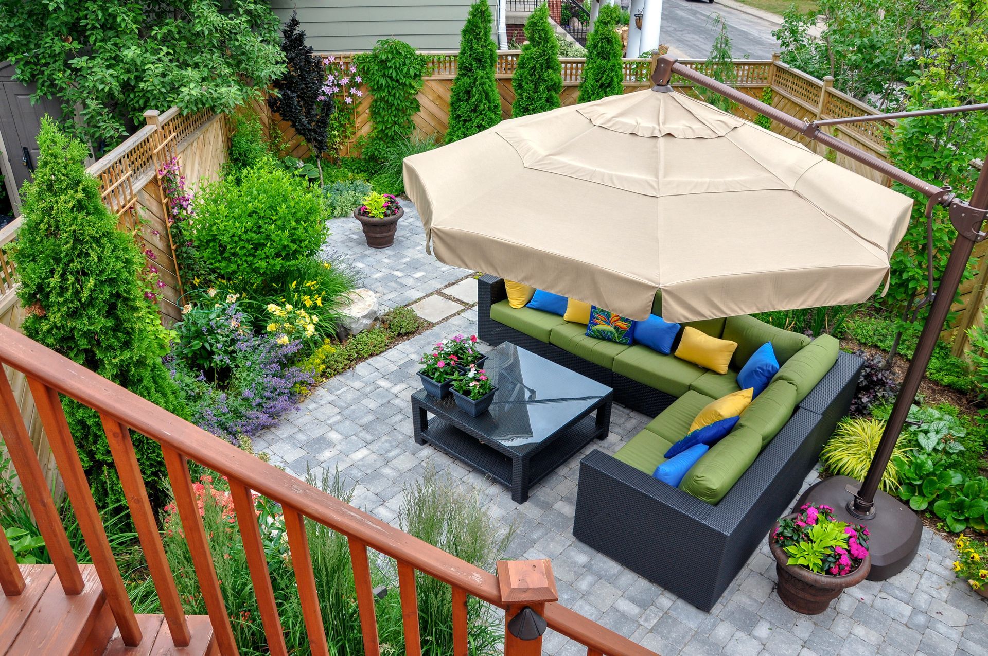 A patio with an L-shaped outdoor sofa, table, and large tan umbrella, nestled in a lush, green, fenced-in backyard.