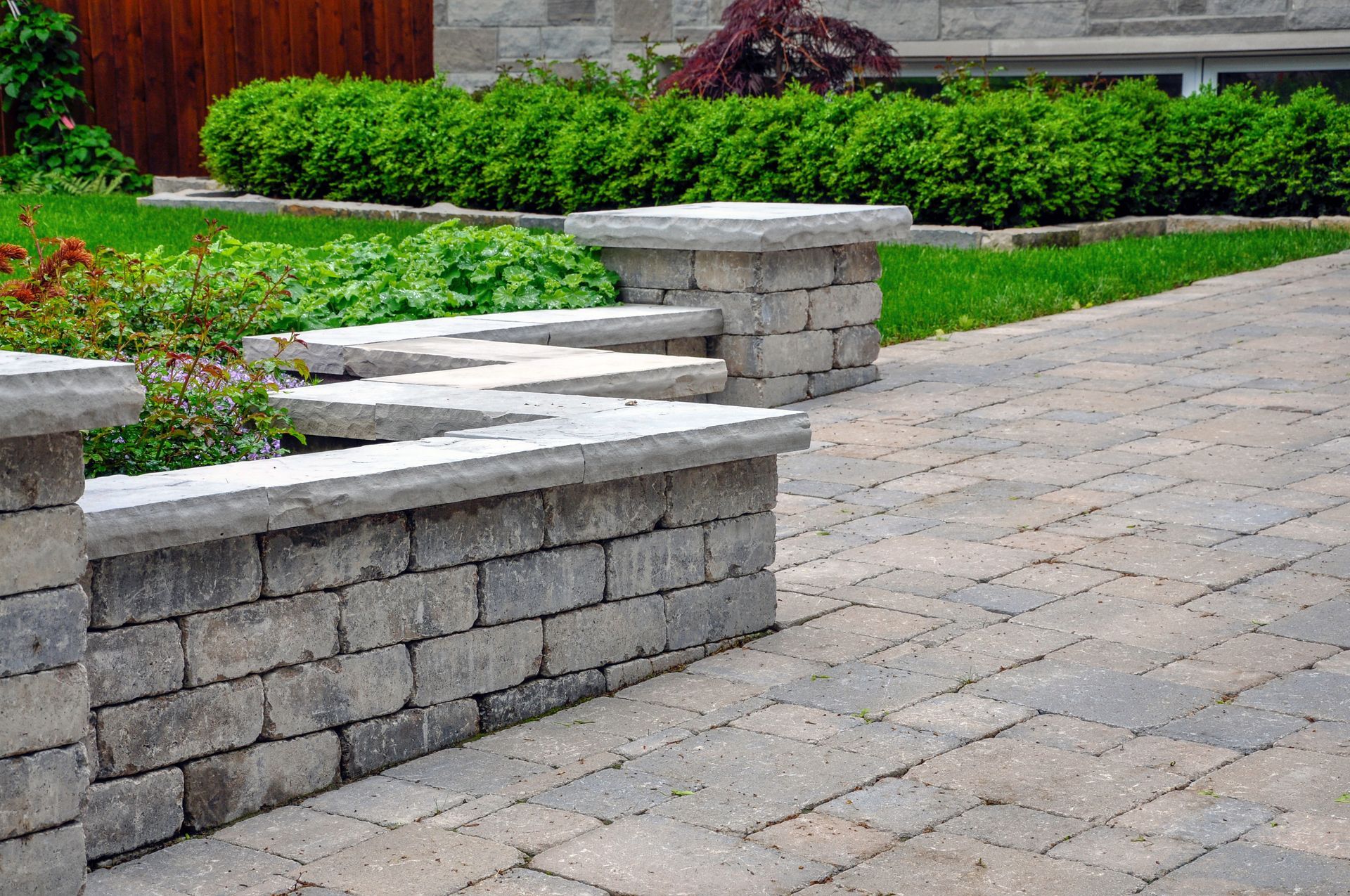 A low, tiered retaining wall built from grey stone bricks, bordering a landscaped garden and paved stone patio.
