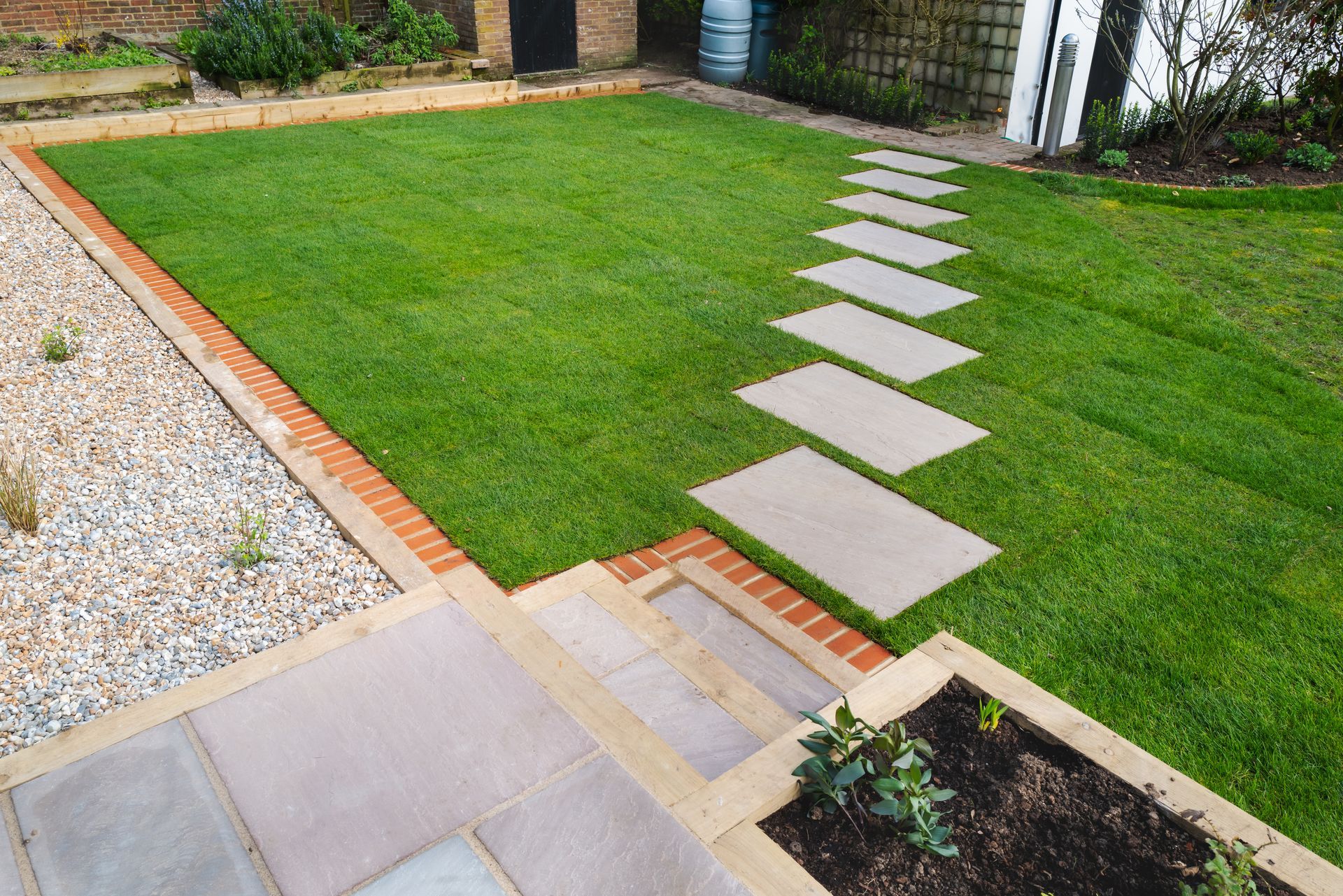 A green lawn with a stepping stone path, bordered by a gravel area and paved steps in a garden setting.