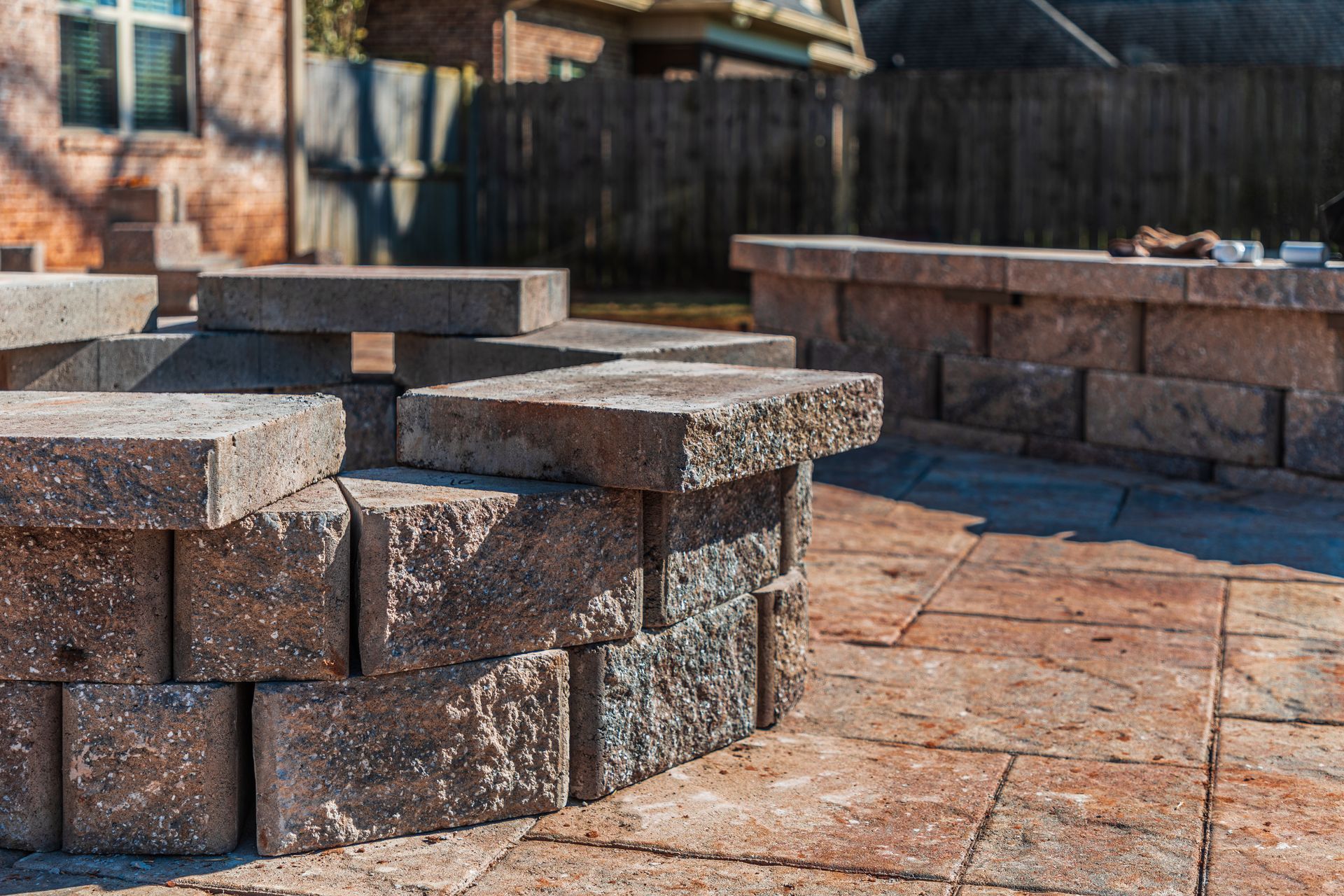 A backyard fire pit and matching seating wall constructed from textured, tan-and-brown stone blocks on a paved patio.