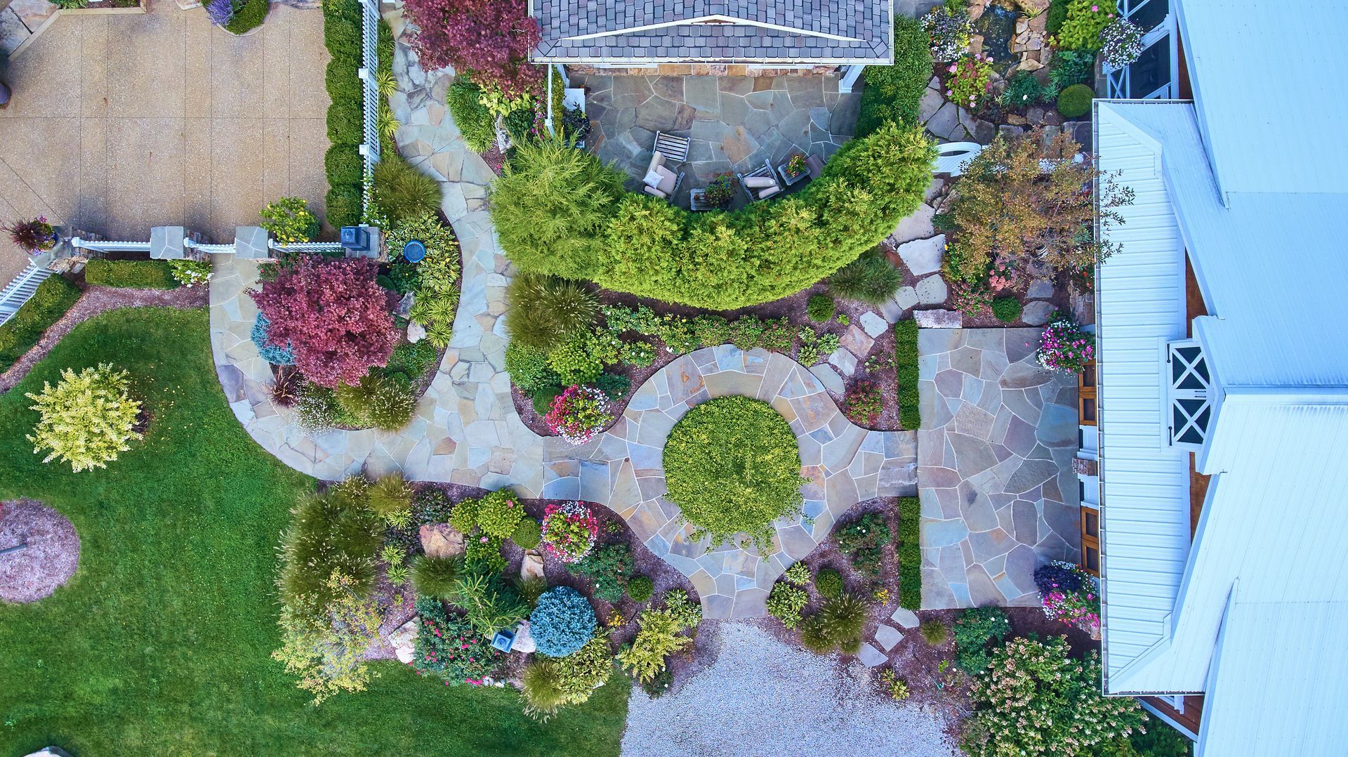 An overhead view of a lush backyard garden featuring flagstone pathways, a circular central bed, and a stone patio.
