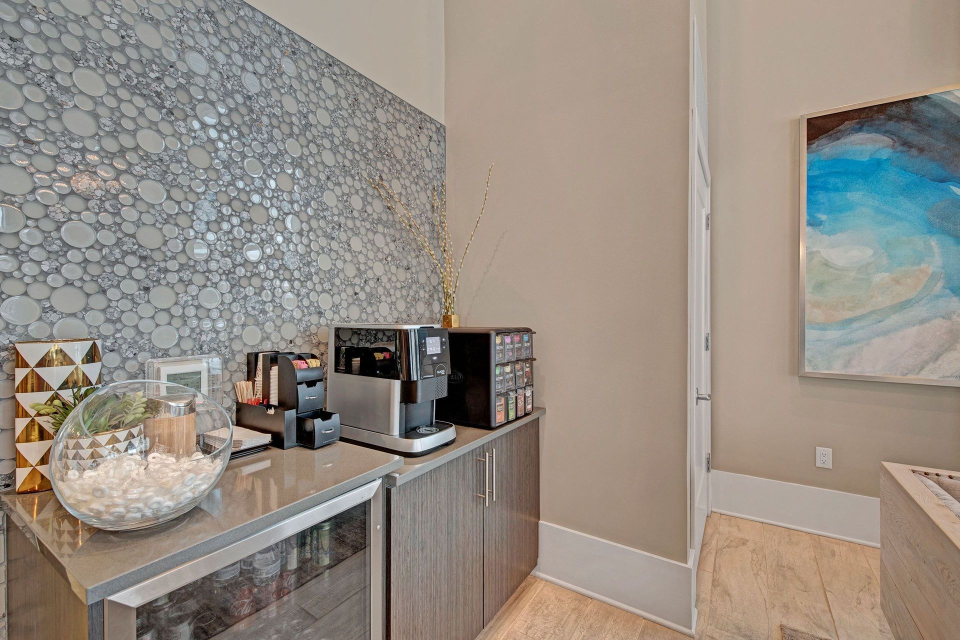 Stylish coffee station with a modern espresso machine, pods, and decor. Bubble-patterned backsplash; abstract blue art on the wall at River House Apartments.