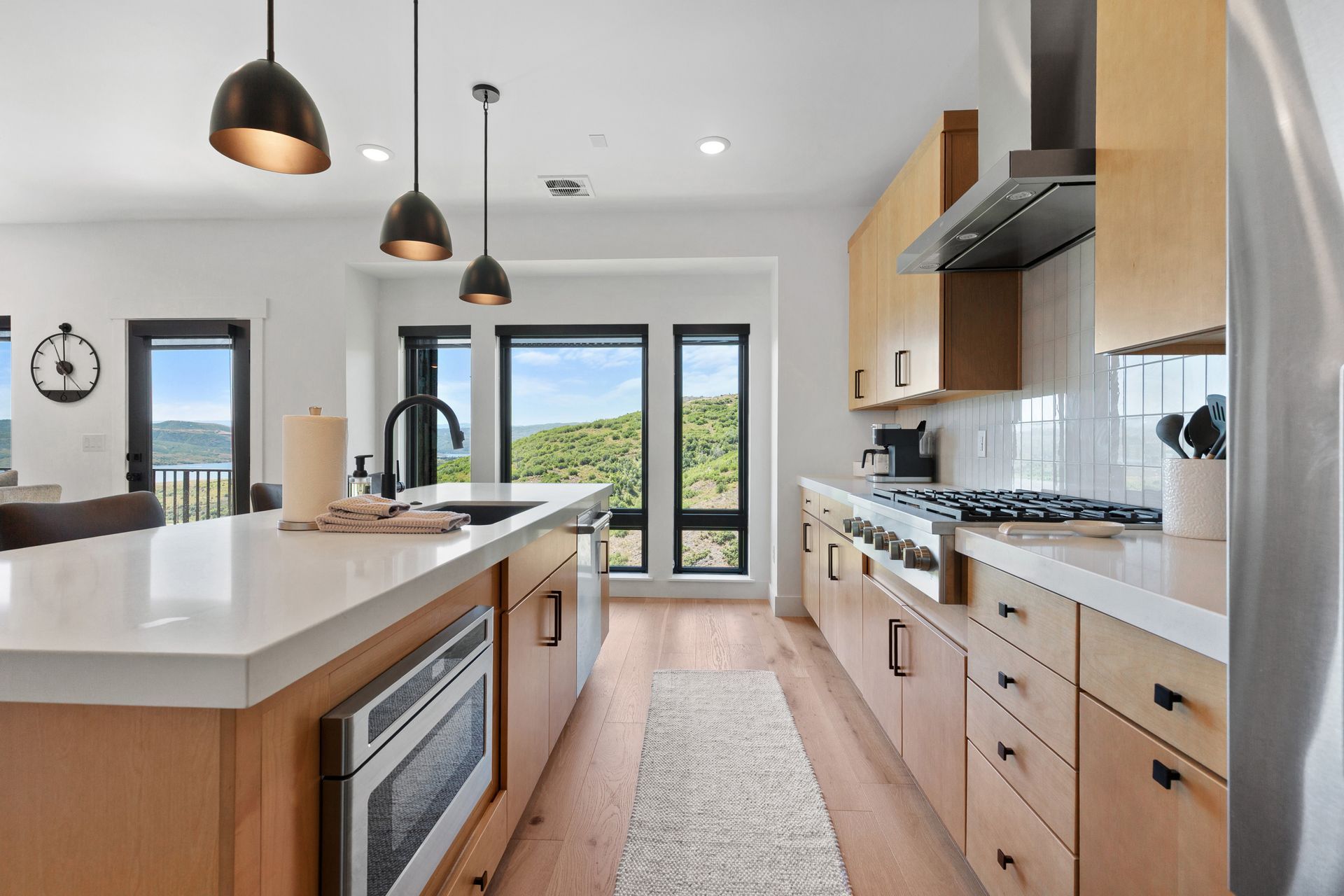 Modern kitchen with wood cabinets, white countertops, stainless steel appliances, and large windows overlooking a scenic view.