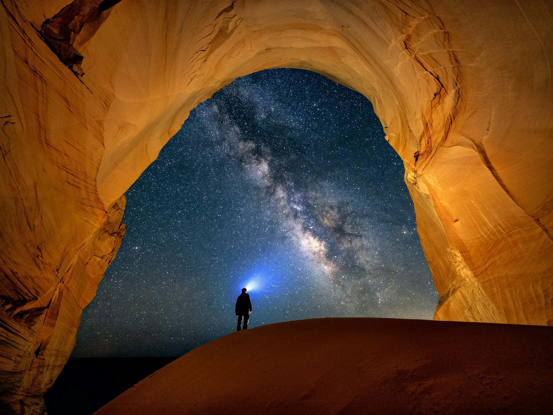 Person with headlamp stands under a rock arch, gazing at the Milky Way.