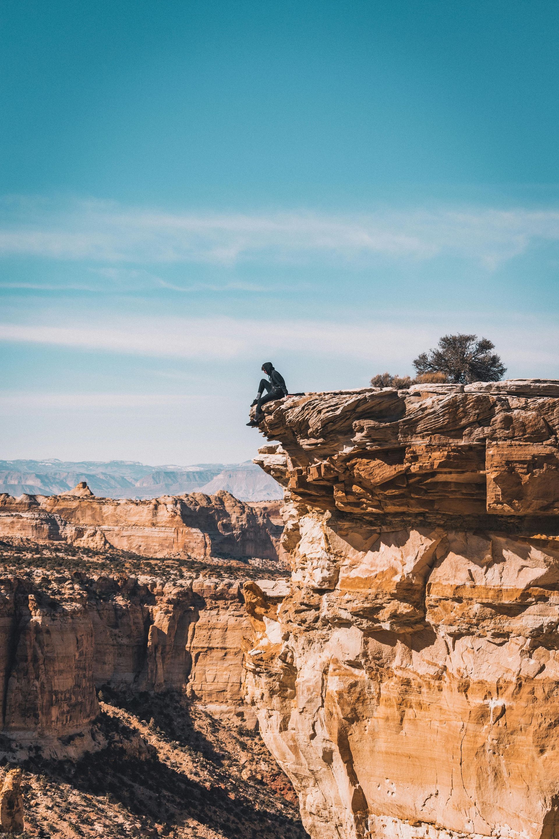 Person sitting on a cliff edge, overlooking a canyon. Blue sky, sunny day.