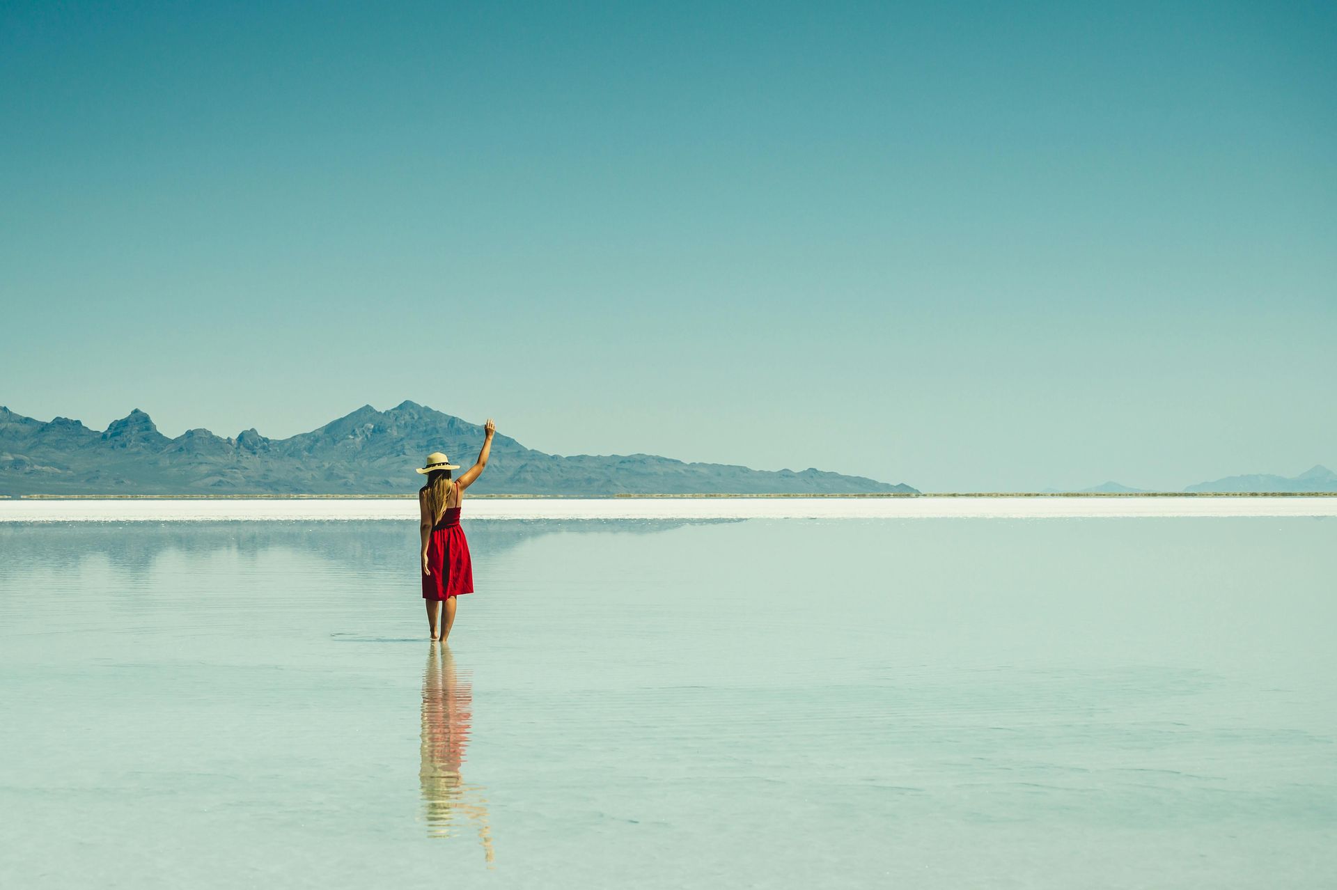 Woman in red dress standing in shallow water, arms raised; vast, reflective landscape under blue sky.
