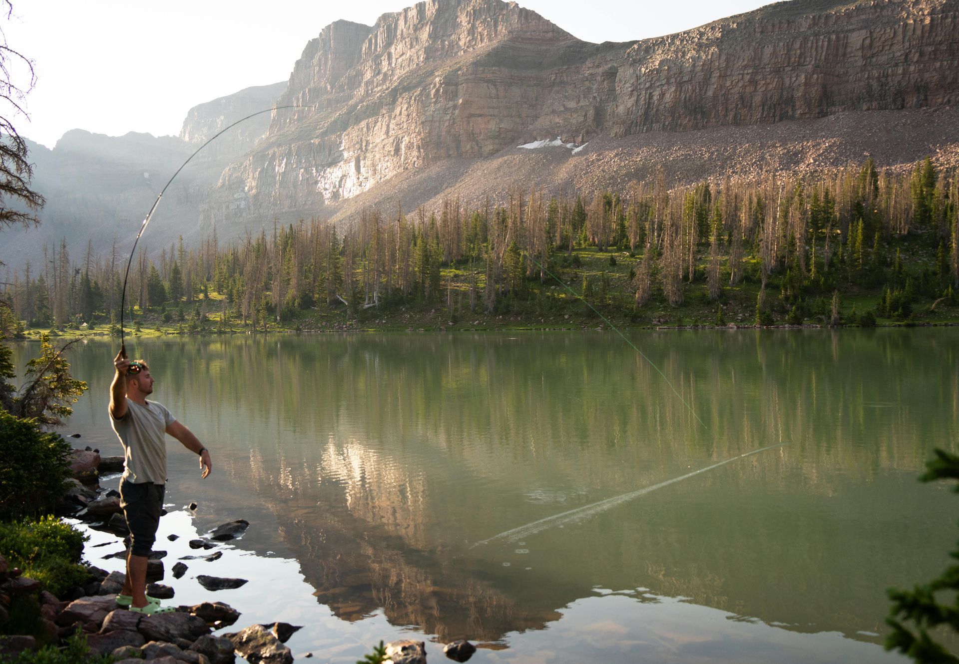Man fly fishing in a serene mountain lake, casting rod with rocky shoreline and mountains in background.