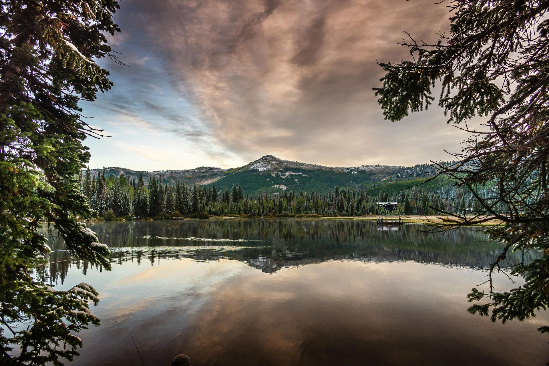 Lake reflects the mountain, trees, and sky. Cloudy, with snowy peaks and evergreen forest framing the view.