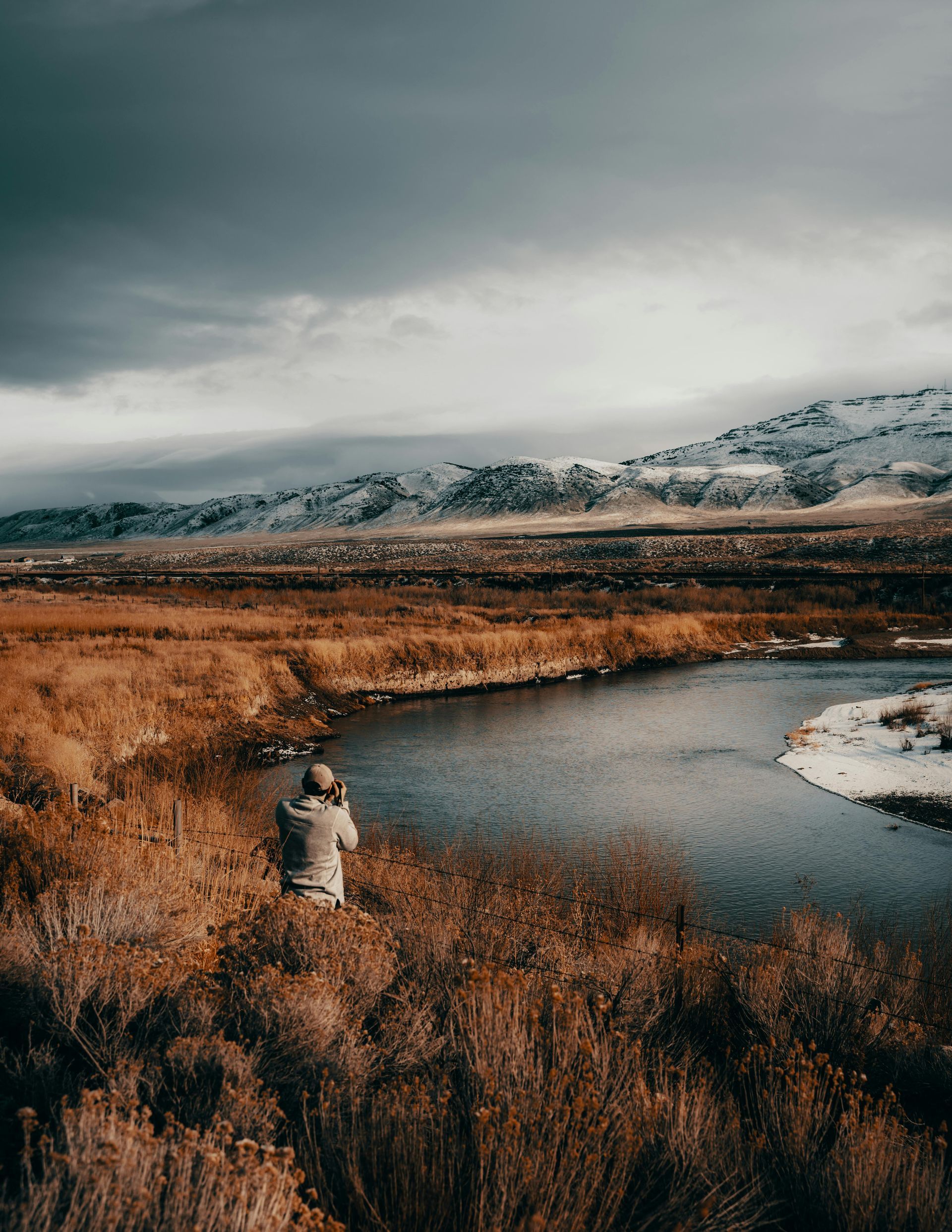 Person photographing a river in a vast, dry landscape under a cloudy sky, snow-capped mountains in the distance.