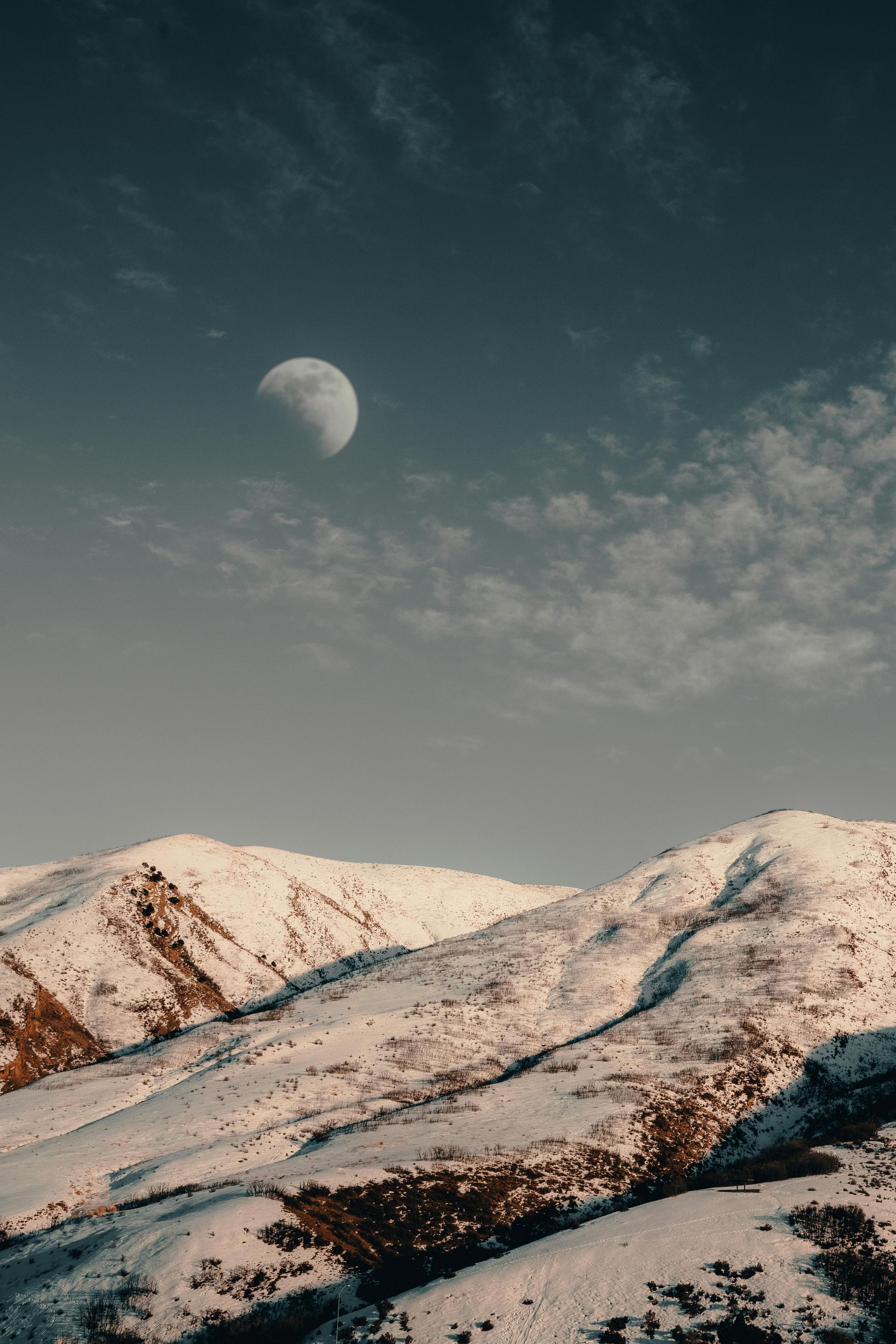 Snow-covered mountains under a dusky sky with a crescent moon and wispy clouds.