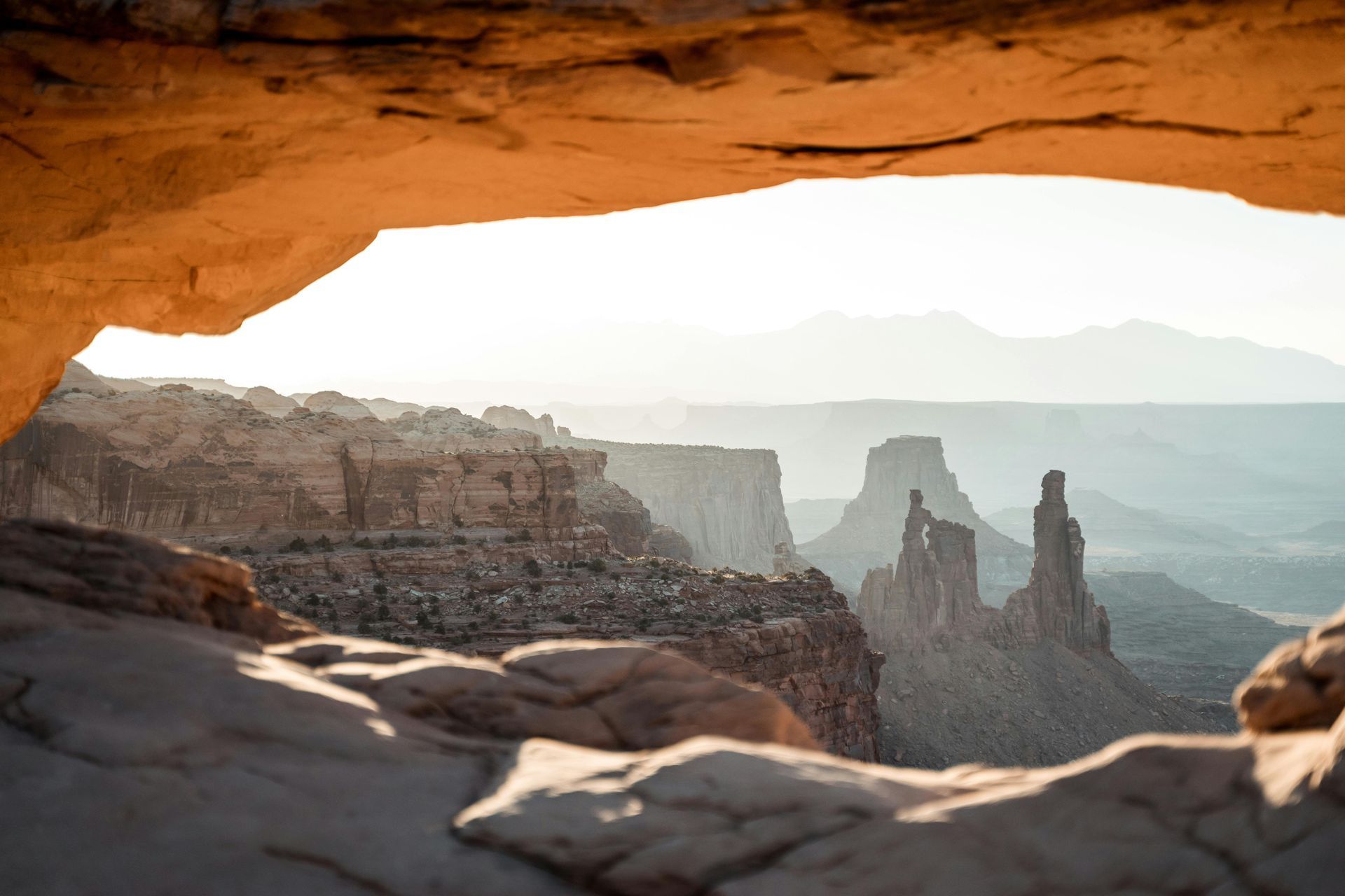 Rocky archway frames a desert canyon view, with mesas and distant mountains in hazy light.