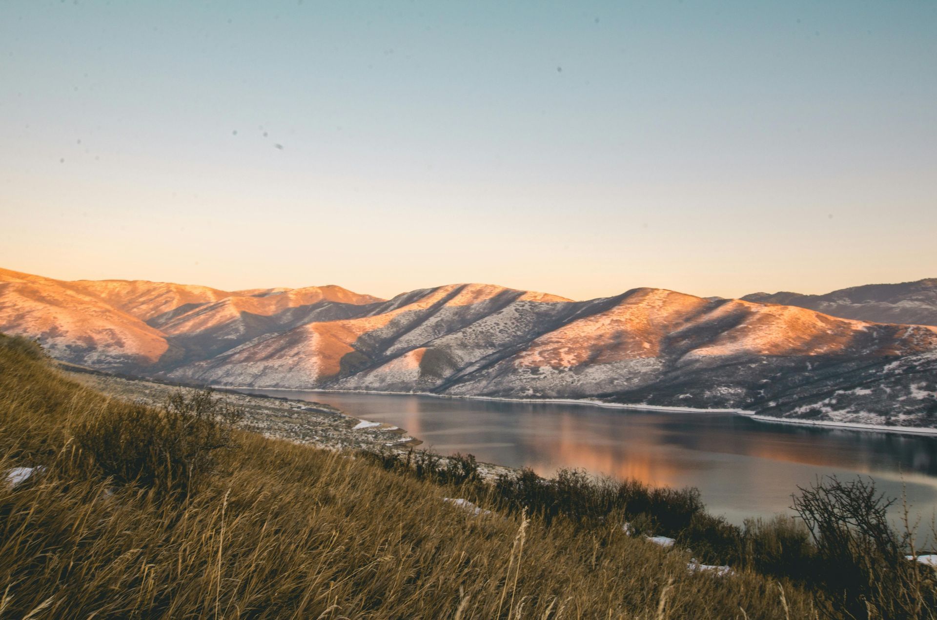 Golden light bathes snowy mountain range reflected on lake under a blue sky.