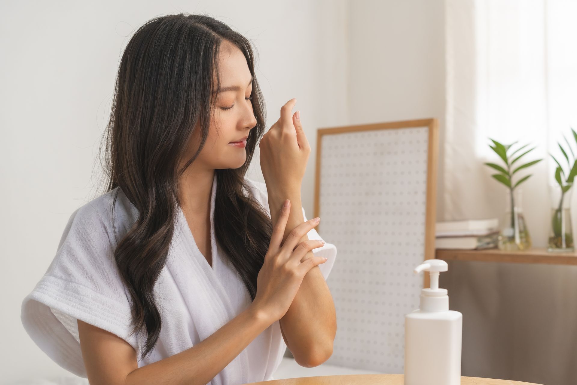 A woman in a white robe is applying lotion to her wrist.