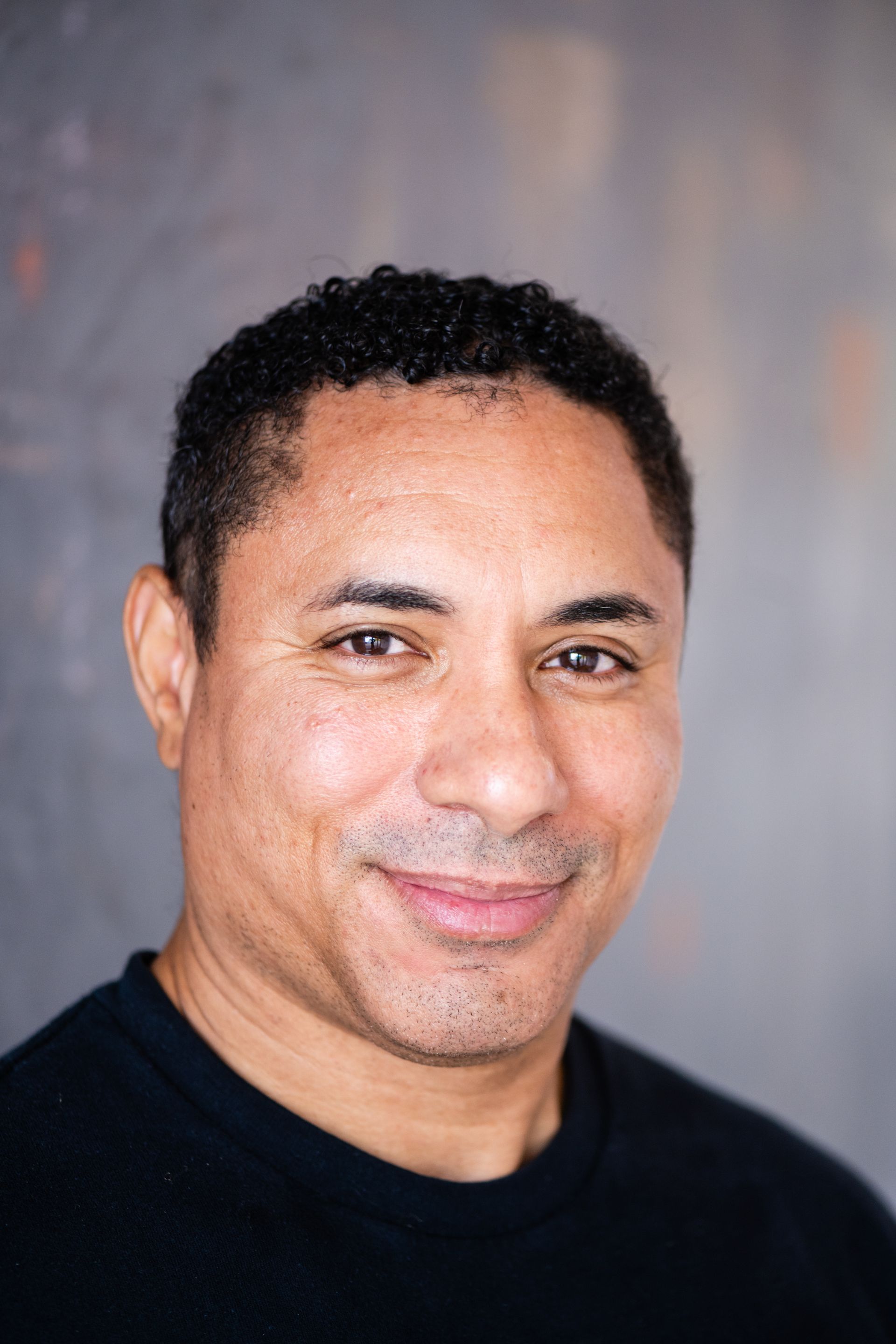 Man with crossed arms, smiling, wearing black scrubs against a gray background.