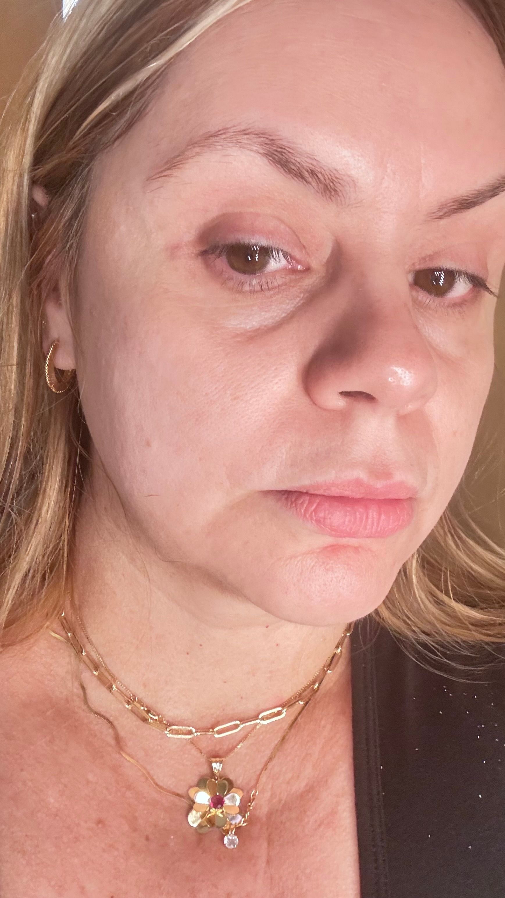 Woman with fair skin, looking down, wearing a gold necklace, small hoop earring, and a dark top.