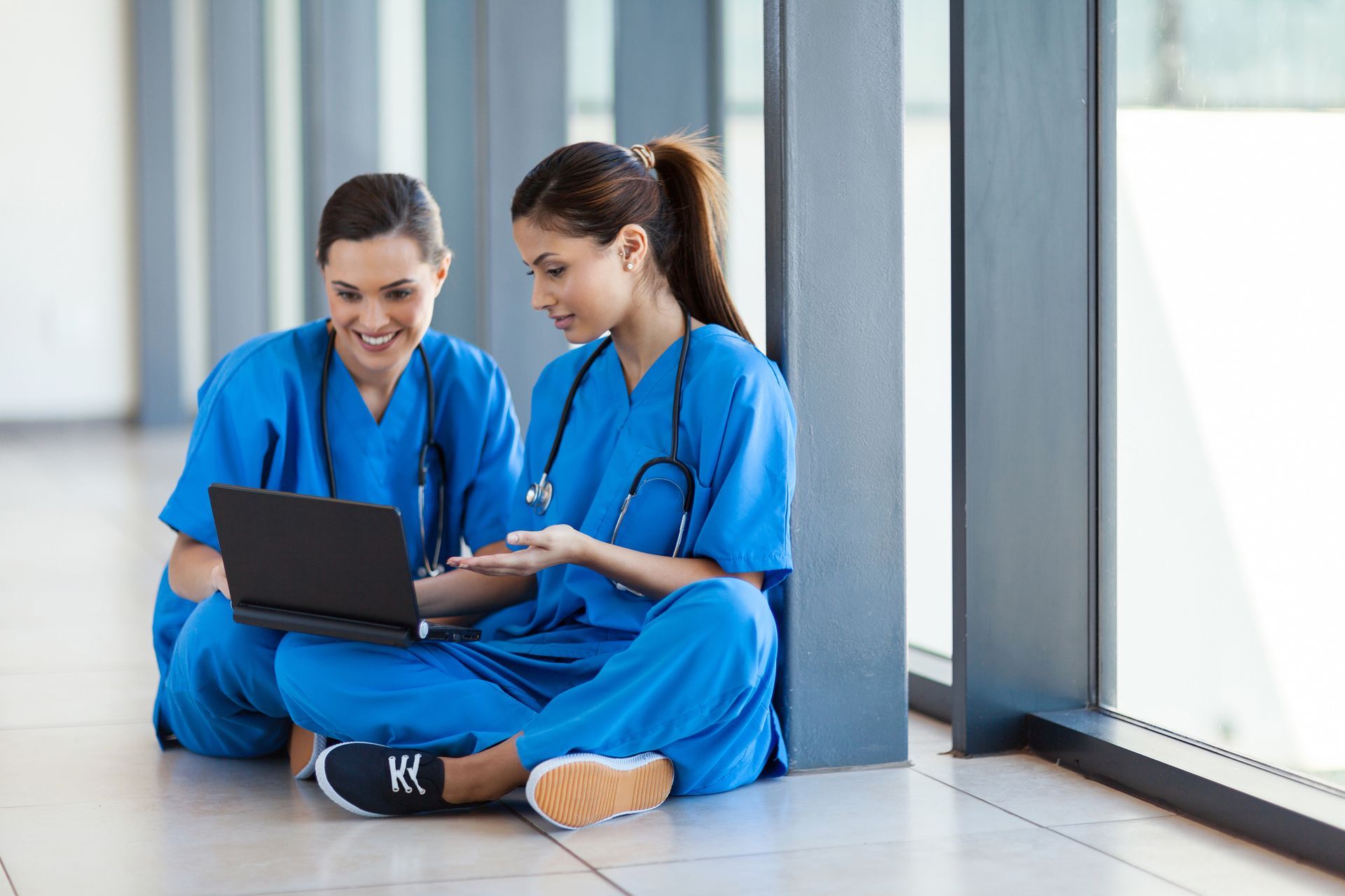 Two nurses are sitting on the floor looking at a laptop.