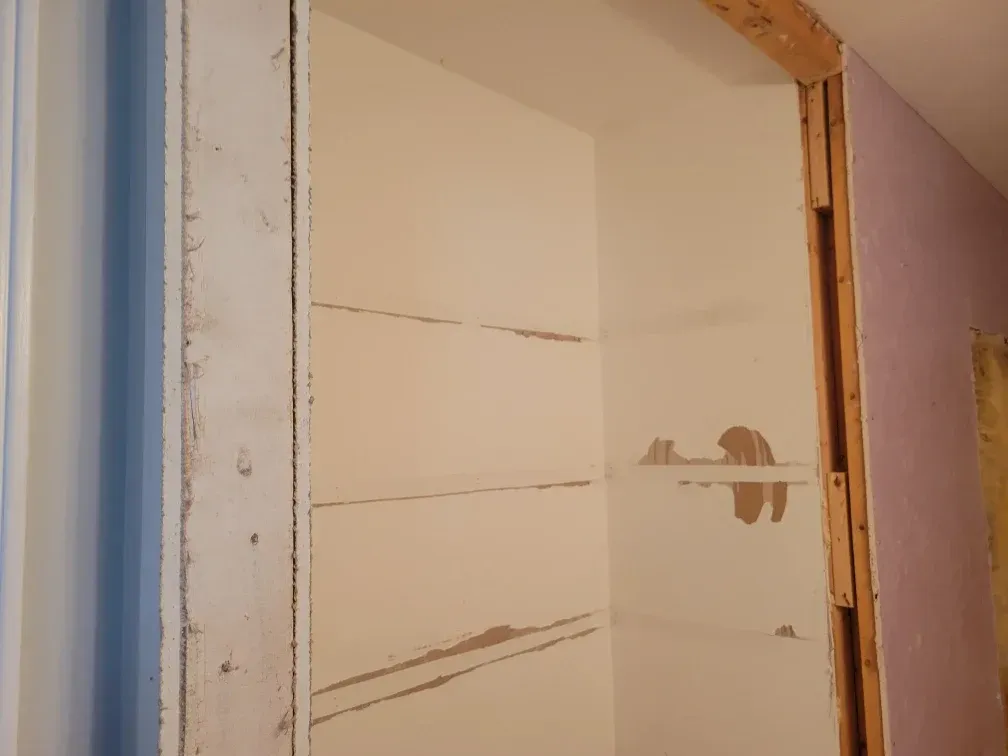 Interior view of an unfinished closet with exposed wood framing and drywall. Light-colored walls, shelves, and a blue door frame are visible.