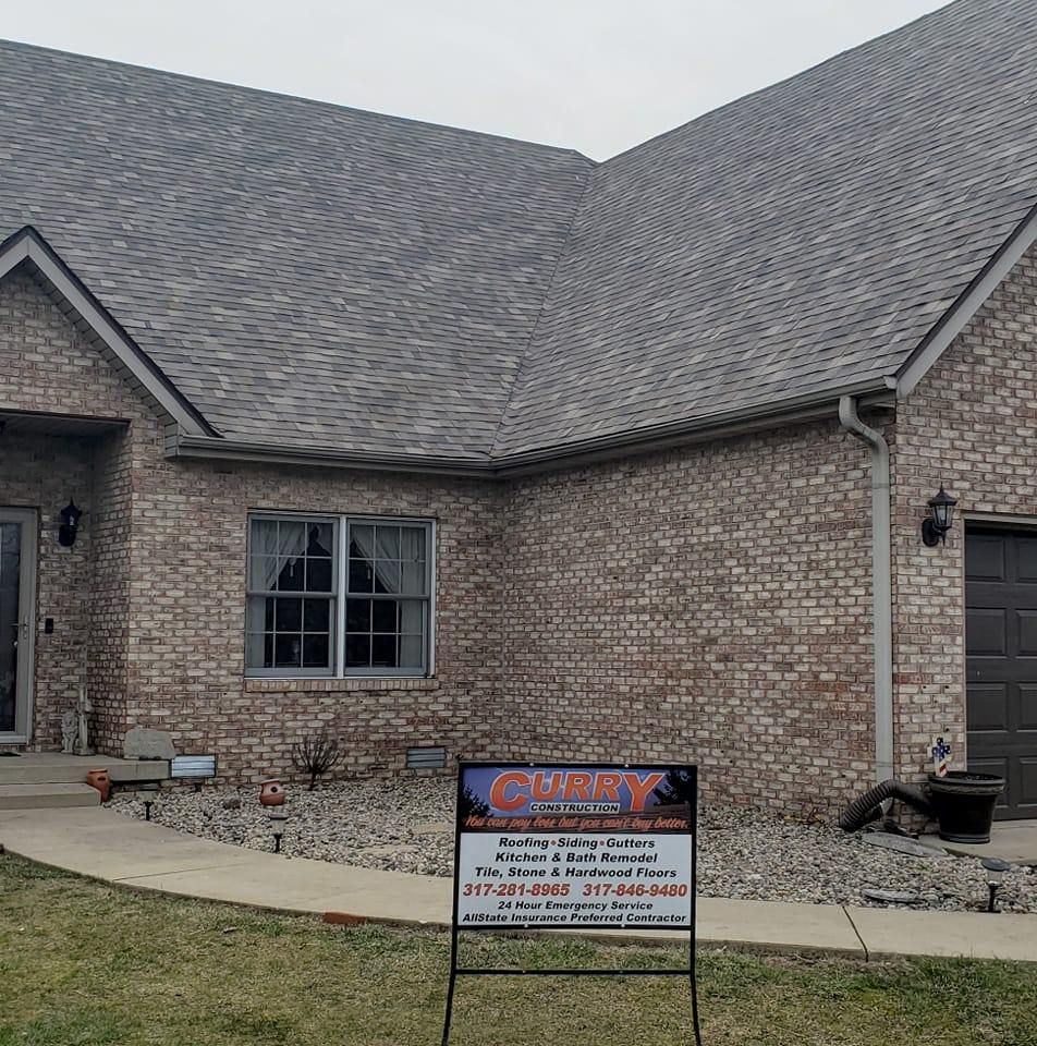 Brick house with gray shingle roof; sign advertising roofing services in yard.
