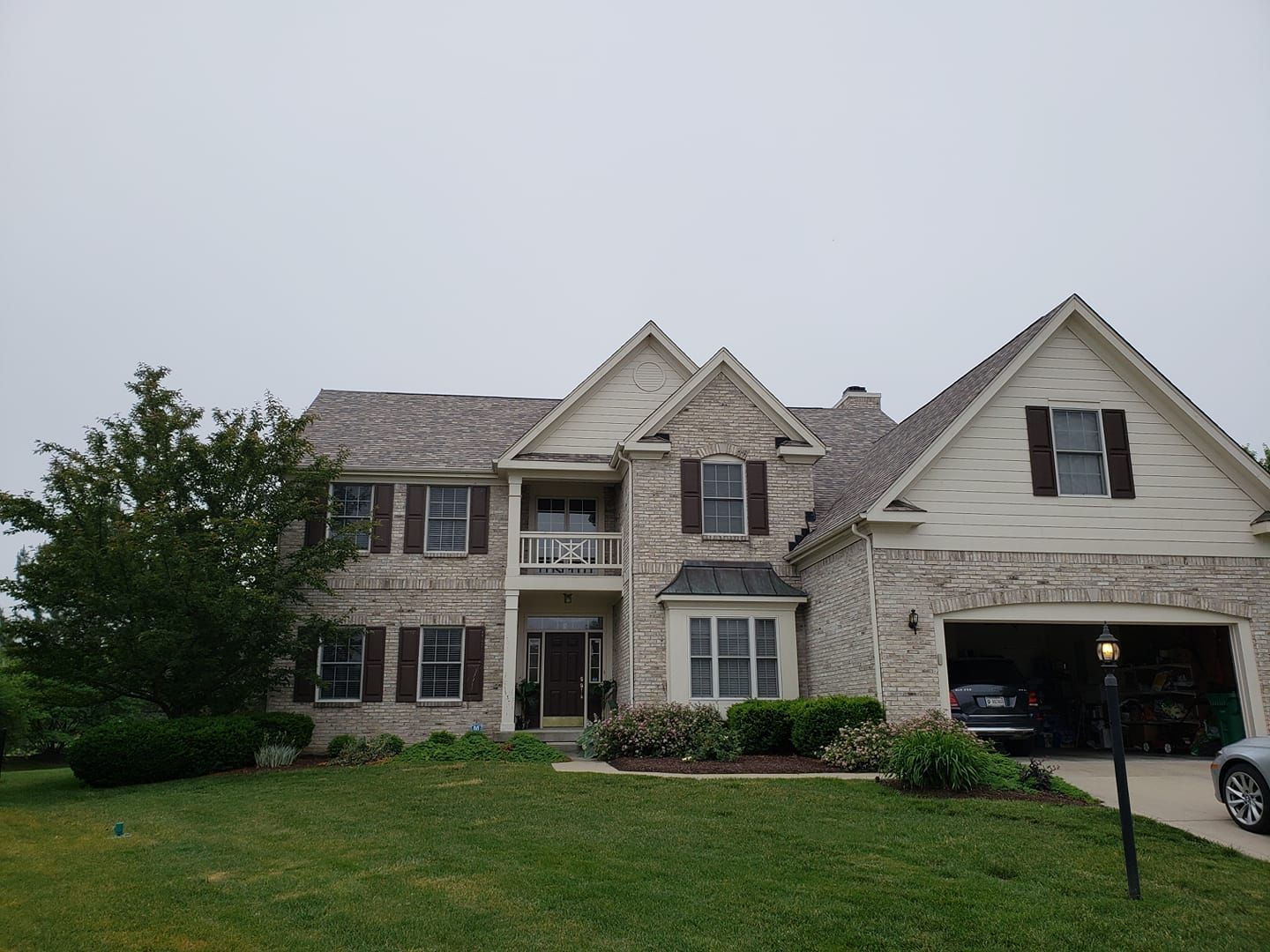 Two-story brick house with brown shutters and garage on a cloudy day. Lush green lawn and tree in foreground.