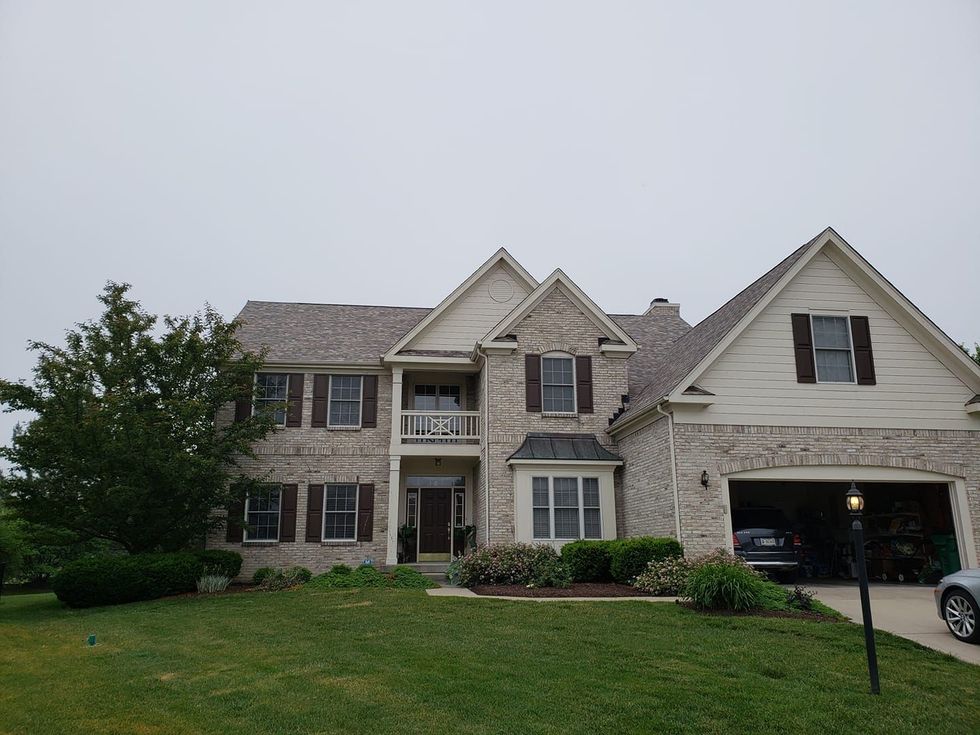 Two-story brick house with brown shutters and garage on a cloudy day. Lush green lawn and tree in foreground.