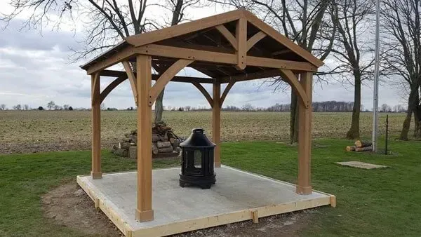 Wooden gazebo on a concrete base, containing a dark object, set in a grassy field.