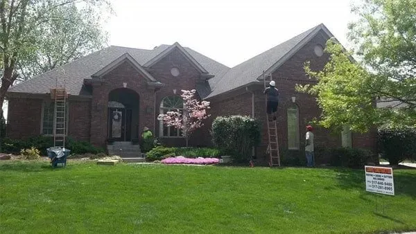 Brick house with workers on ladders; yard with sign; green grass and trees.
